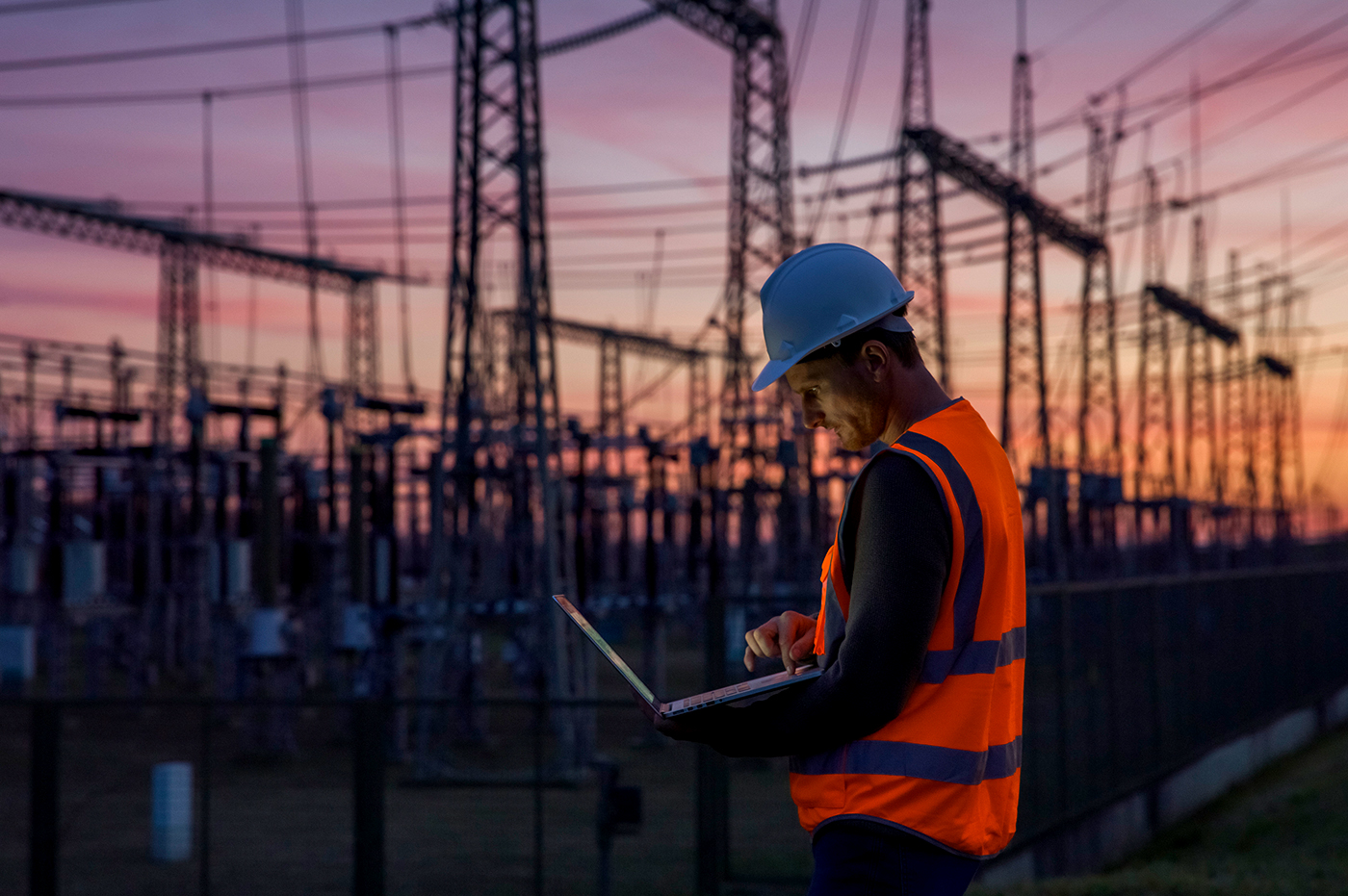 A man in vest and hard hat working on his laptop at an energy plant.