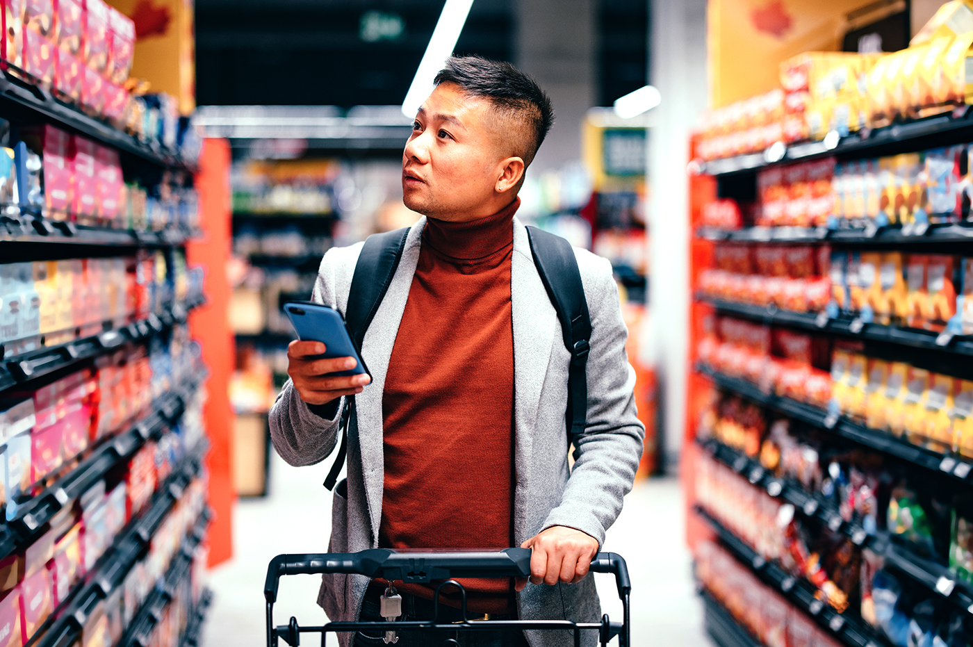 Man shopping in a grocery aisle pushing a shopping cart with a phone in his hand