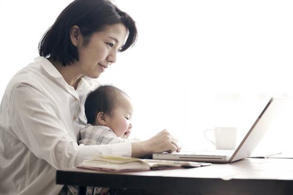 A woman working on a laptop and holding her baby.