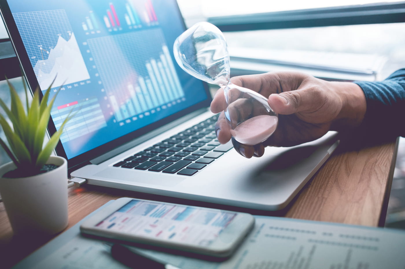 Male Hand Holding Sand Hourglass Next to Smart Phone and Laptop