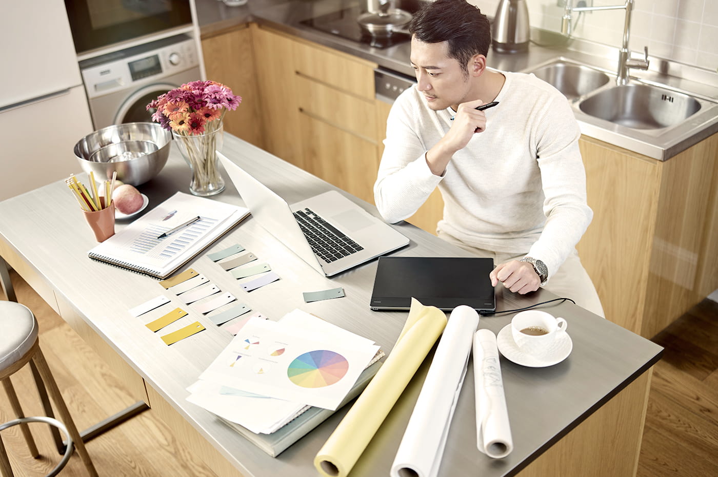 Man sitting at a counter working on laptop and looking at design materials.