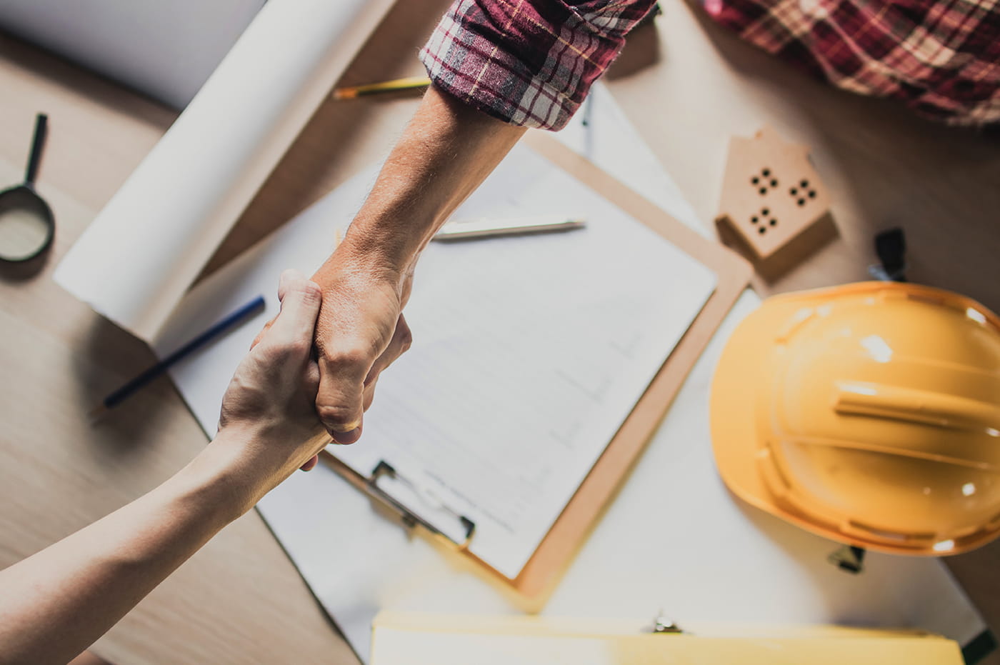 Two people shaking hands over construction helmet and materials.