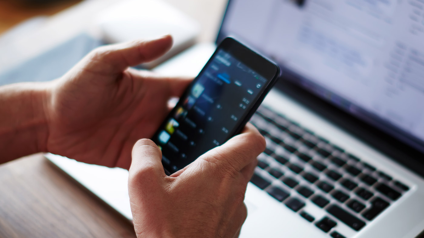 Hands typing on cell phone with a laptop in background.