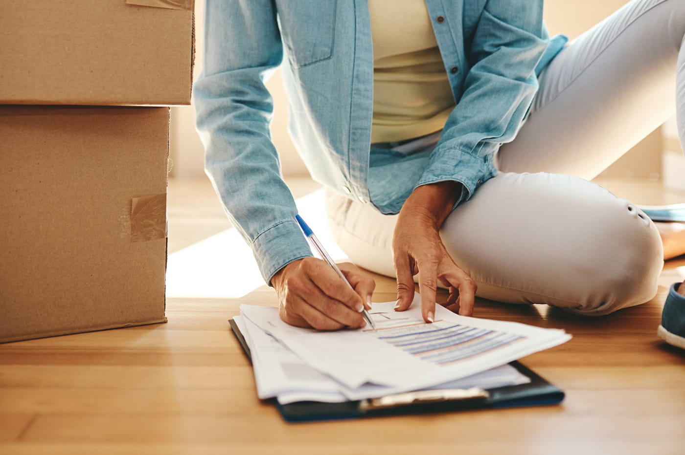 Person sitting on floor writing on papers next to moving boxes.