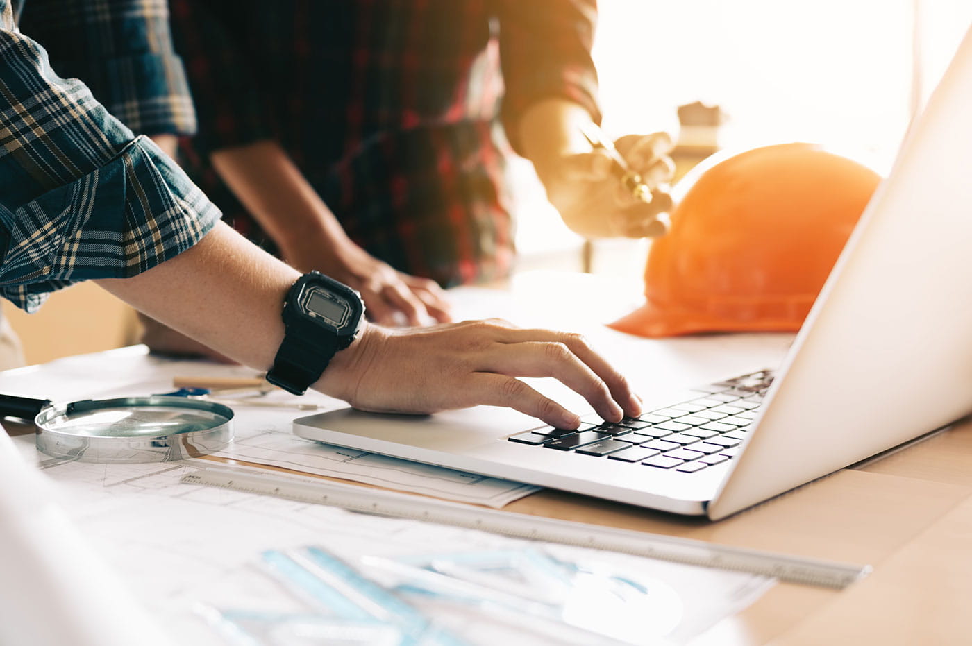 Two men in flannel shirts working on a laptop with a hard hat next to the computer.