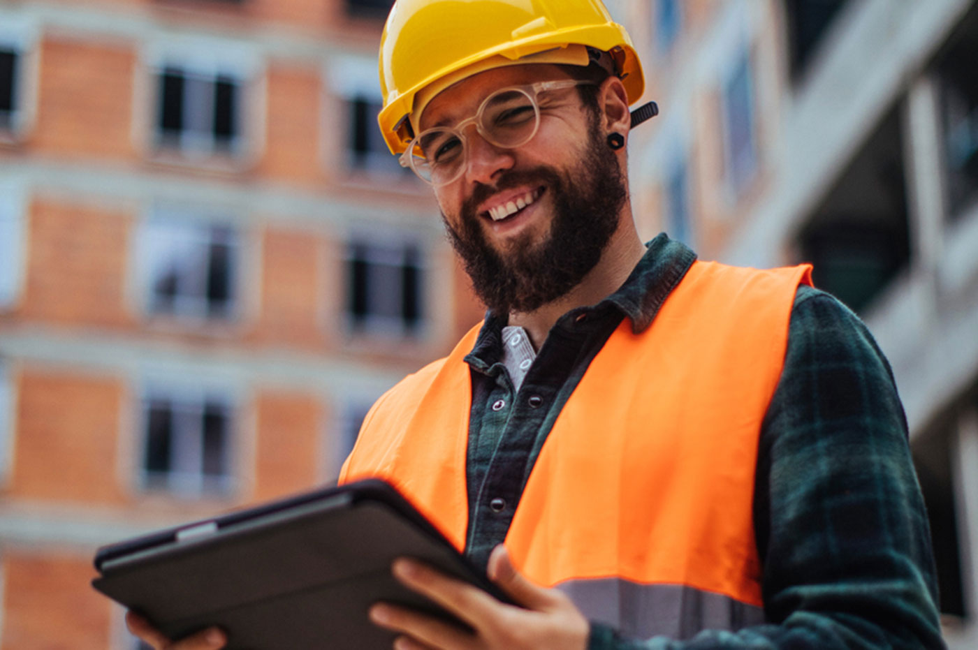 A man in a hard hat, safety vest, and glasses smiling at his tablet while he works.
