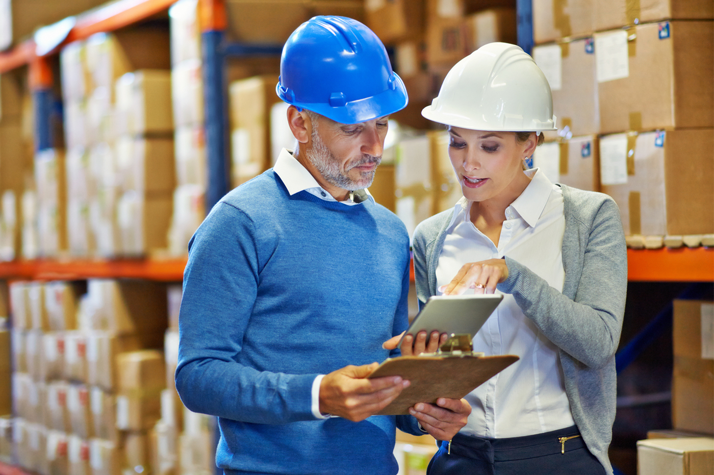 A man and woman with construction helmets on in a warehouse looking at a tablet device. 