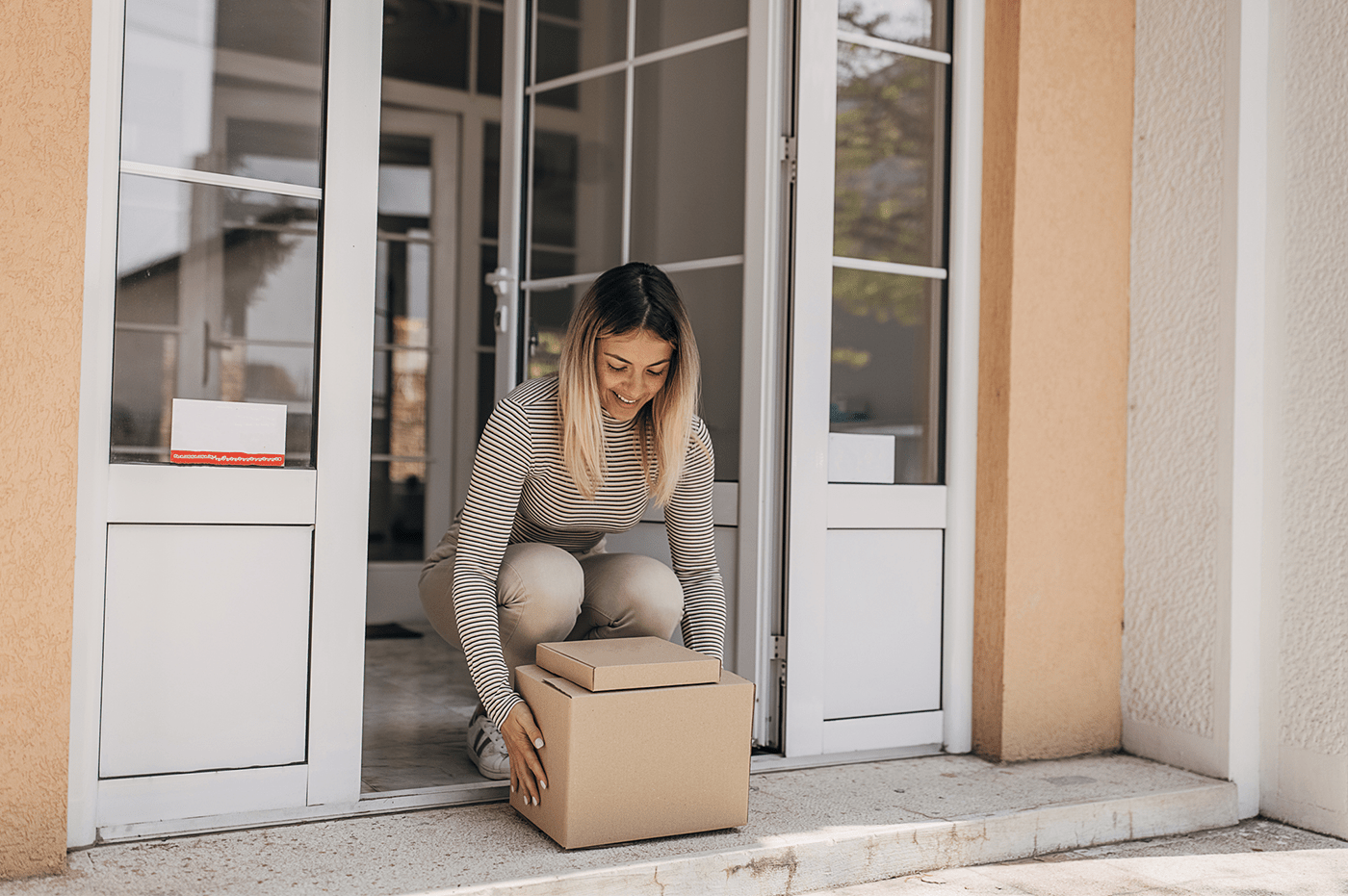 A woman picking up a package in front of her house on her porch. 