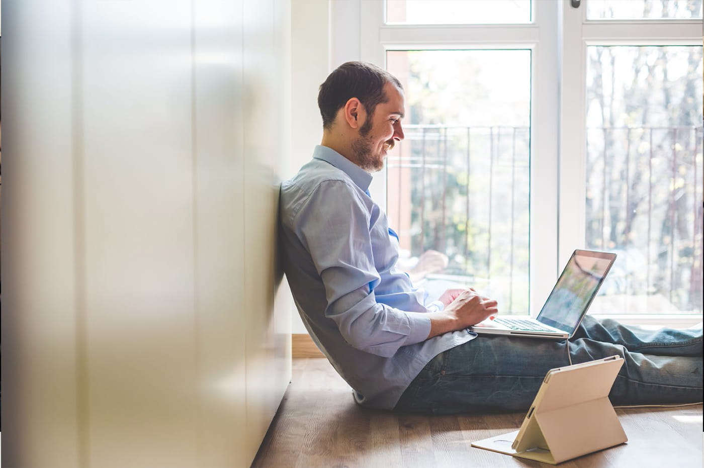 A man sitting near a window working on a laptop and tablet.