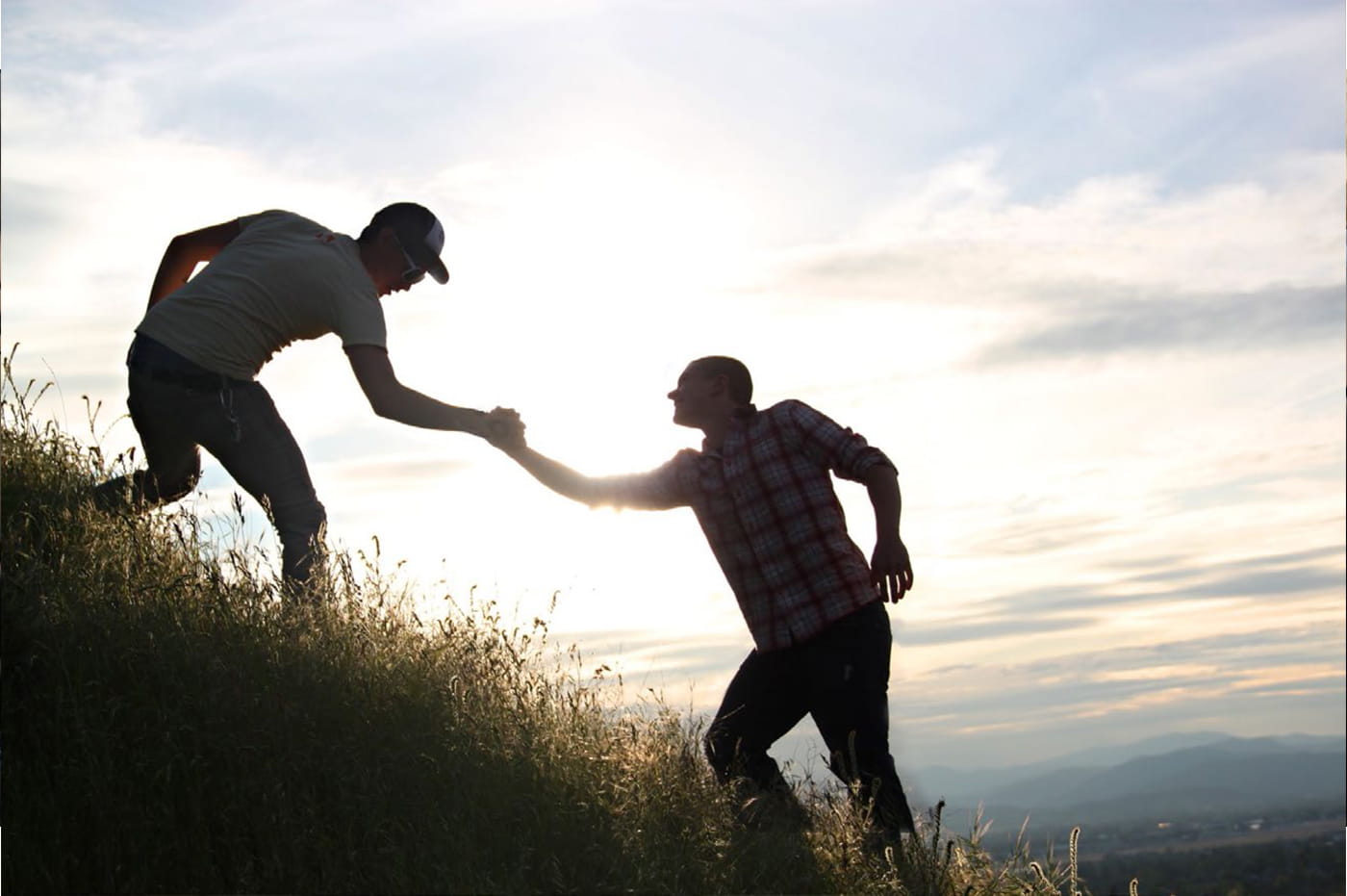 Two men helping each other climb up a steep hill.