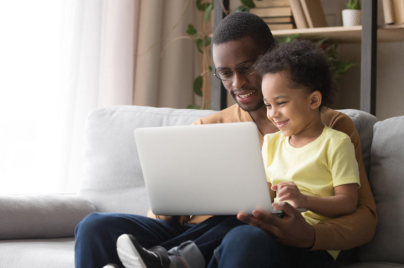 An African-American father and son sitting on the couch and looking at a laptop together.