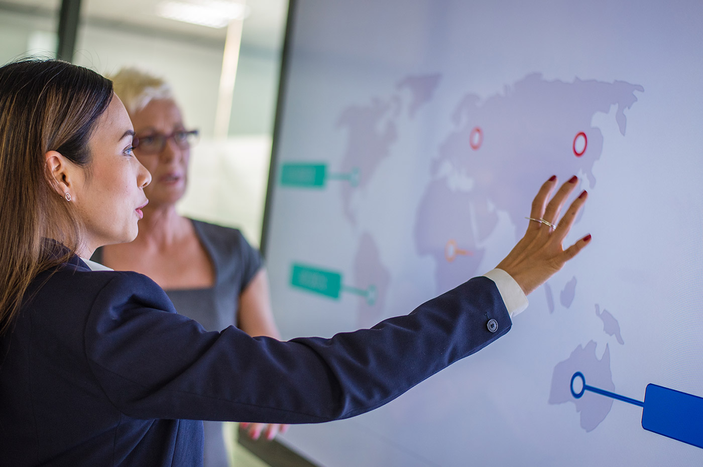 Two business women looking at a world map on a large screen and discussing.