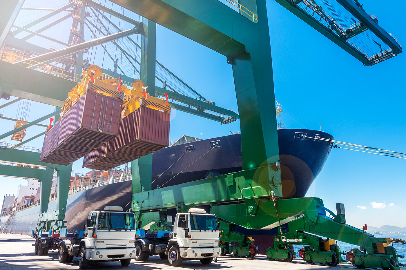 Two large shipping crates being lifted onto a shipping boat by cranes.