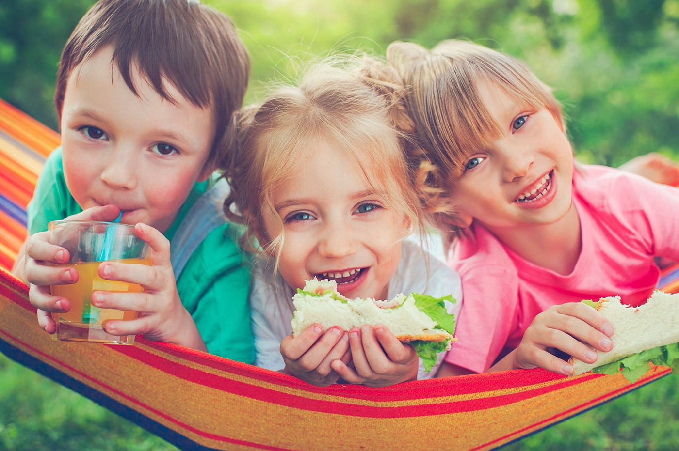 Three children sitting together in a hammock eating sandwiches.