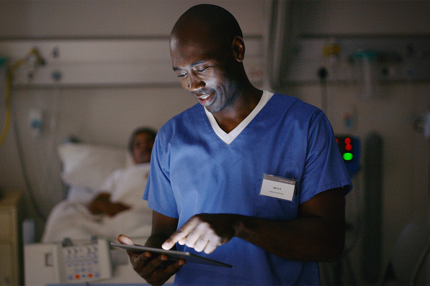 A male doctor looking a tablet with a patient in the background in a hospital room. 
