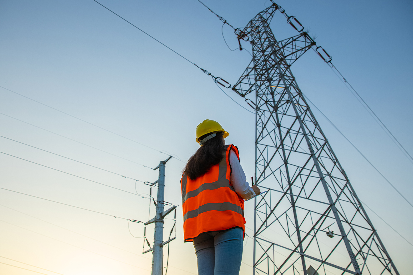 Woman standing in front of transformer outside in a hard hat and construction vest taking notes