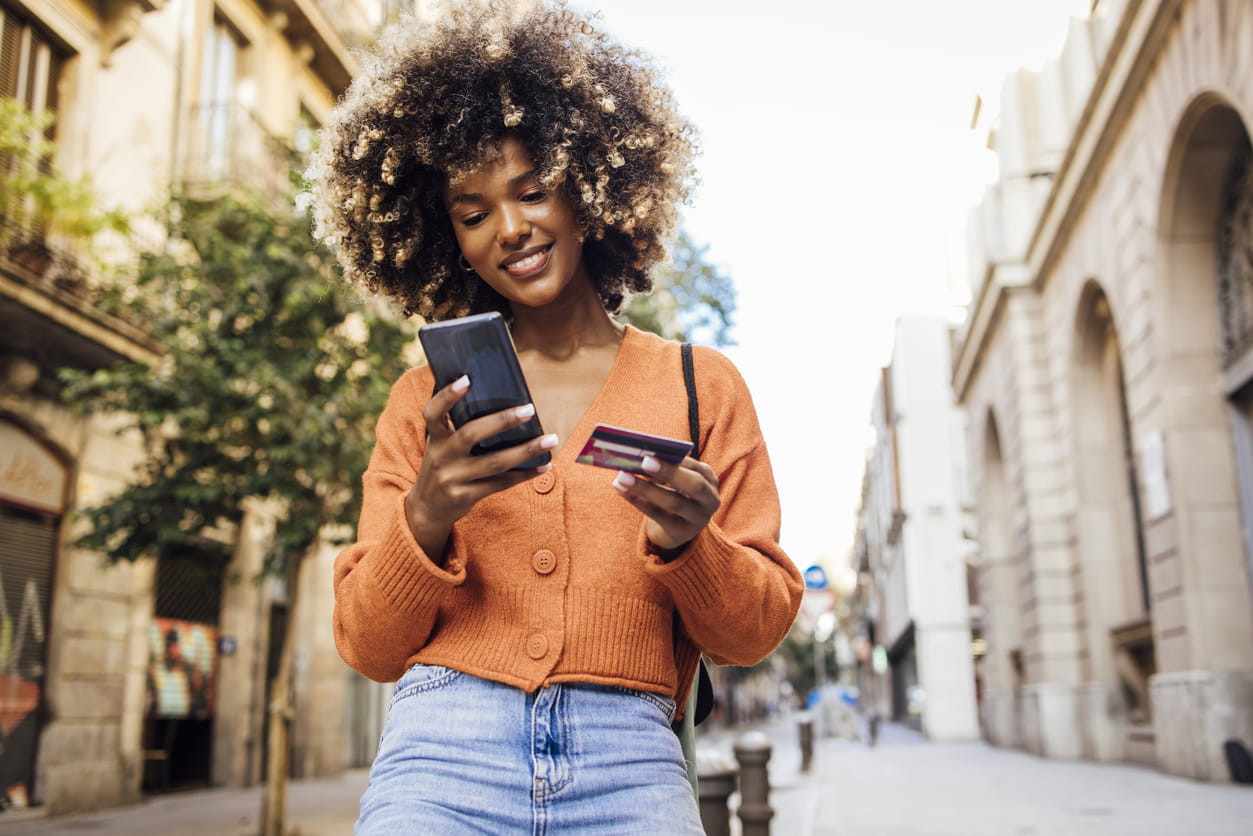 Woman standing in street looking at phone holding credit card