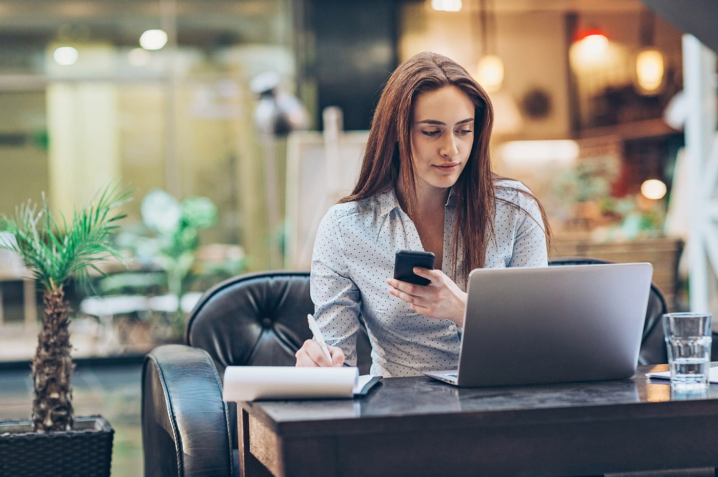 A woman working on a laptop and cellphone while taking notes.