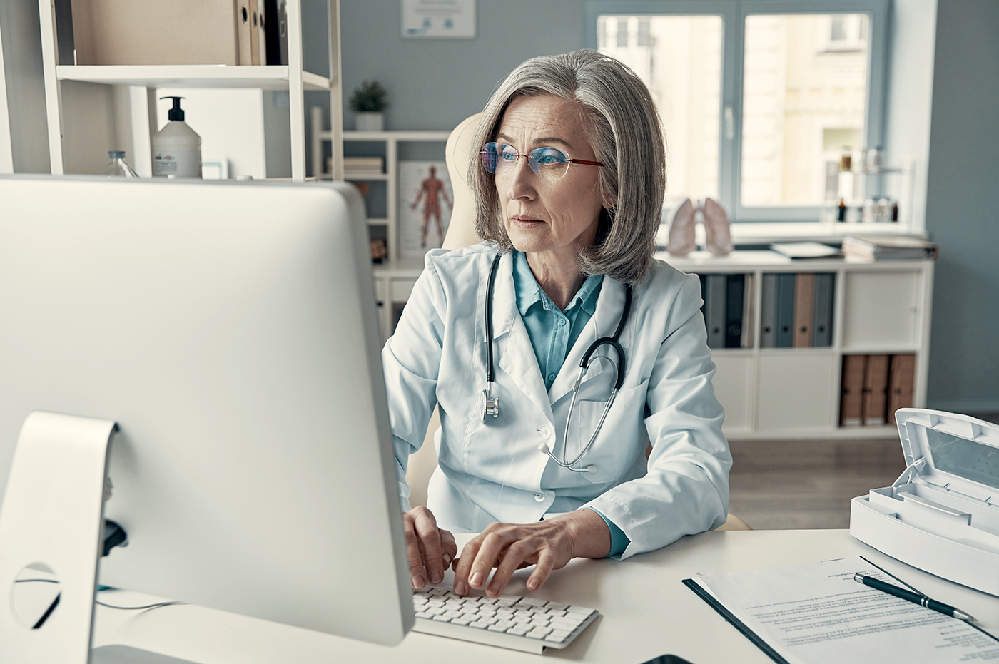 An older female doctor working on a desktop computer.