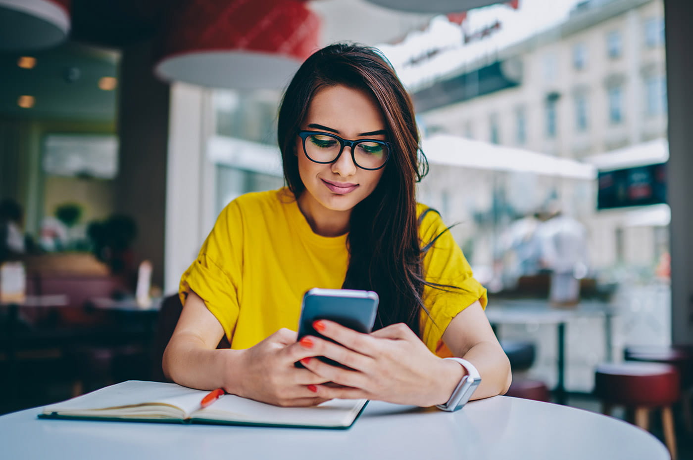 A young woman with glasses sitting at a table looking at her cellphone.