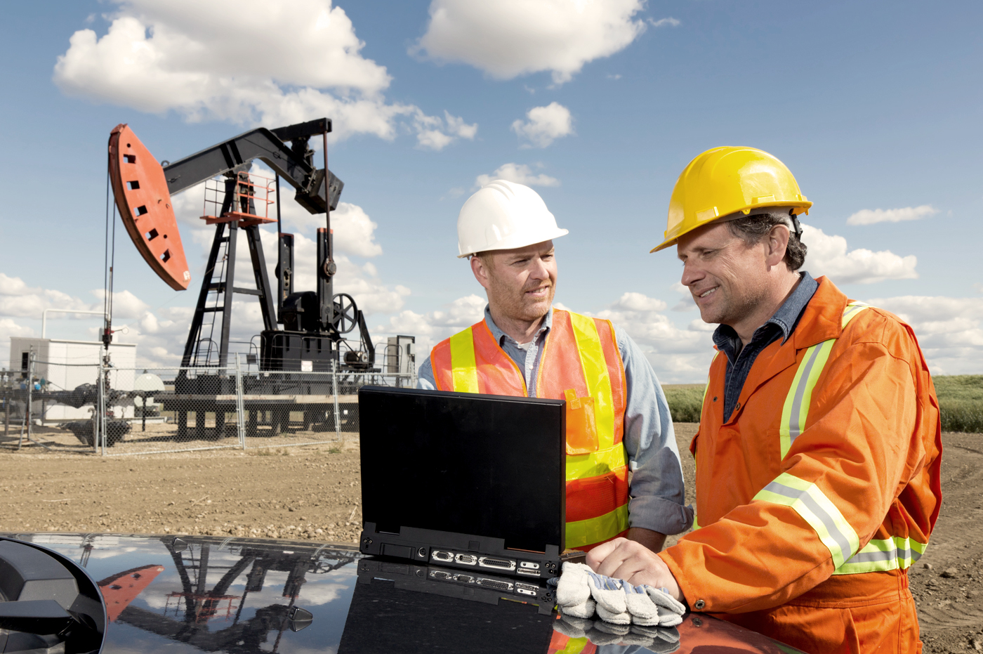 Two men in hard hats and safety vests working on a laptop at a drilling site.