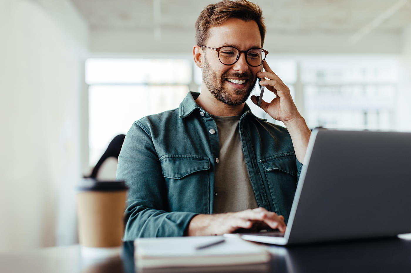 Man smiling on the phone while sitting down at a table and typing on a laptop