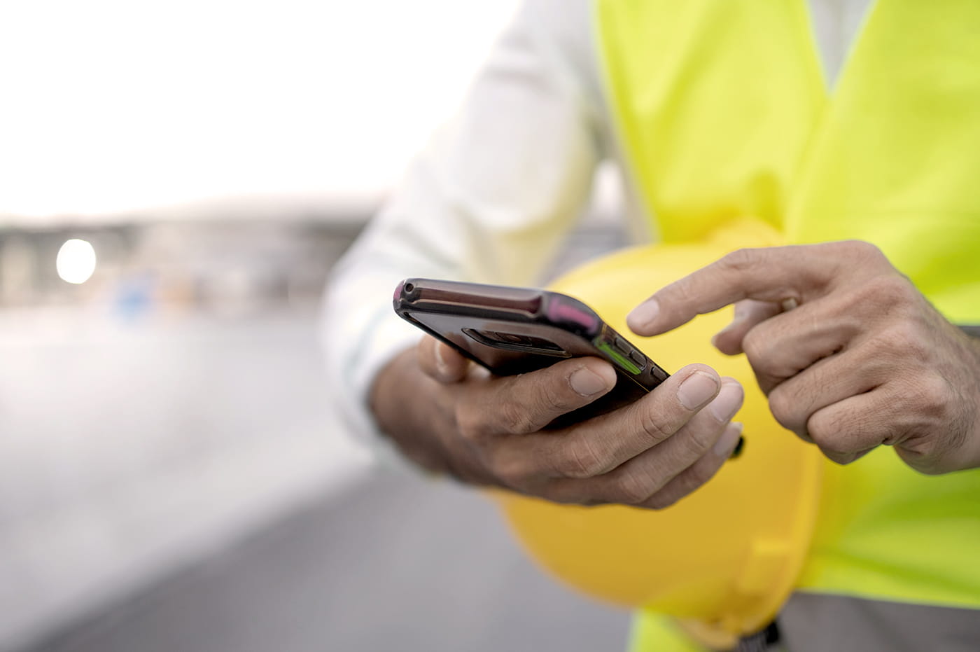 A construction worker holding a helmet under there arm while looking at a cell phone in their hands.