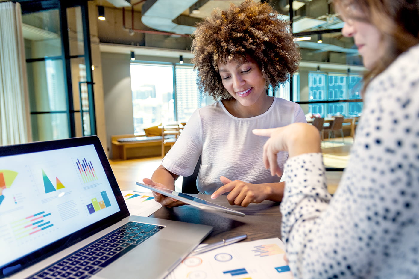 Two businesswomen looking at a laptop with charts and also looking at a tablet. 