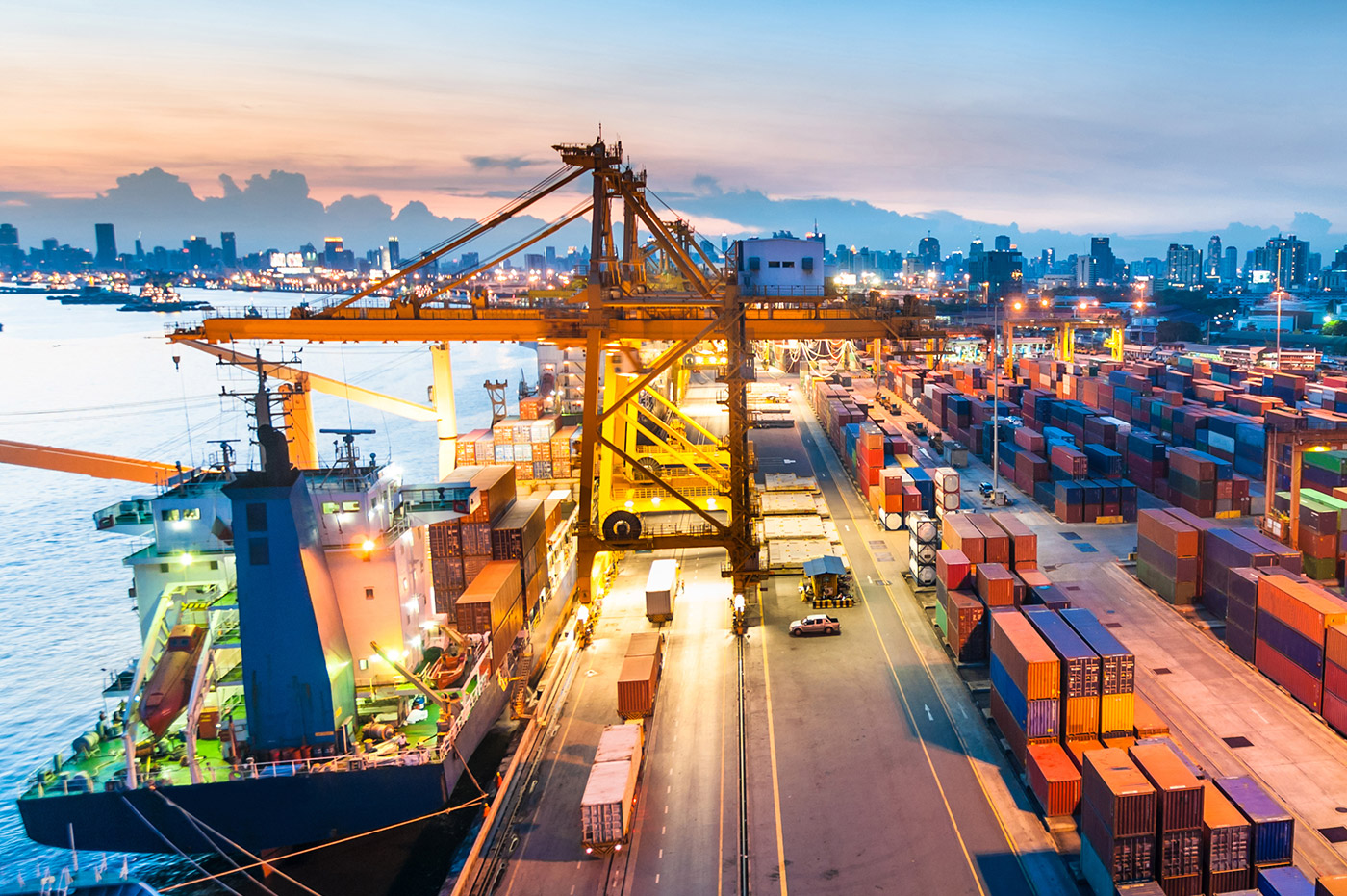 View of a shipyard with a ship, shipping containers, and parked cars.