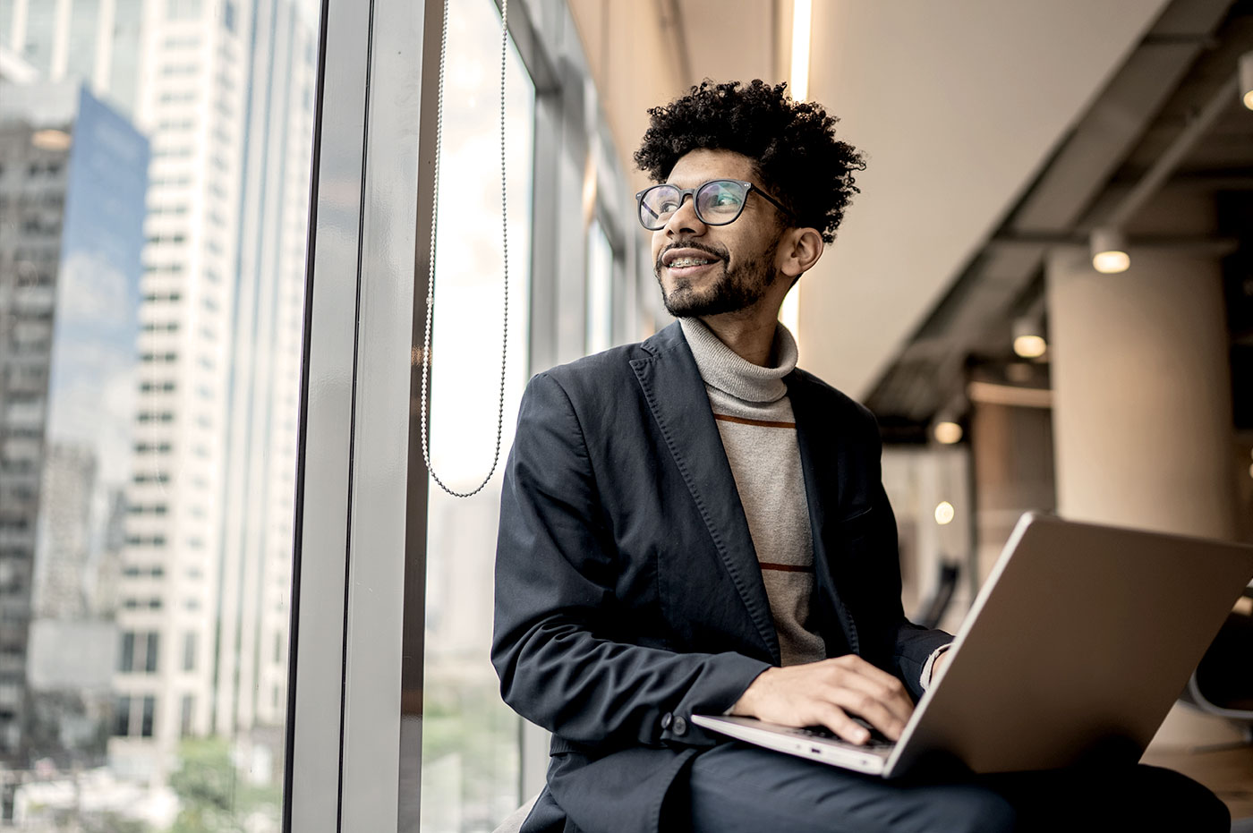 Man working on laptop and sitting in a window smiling.