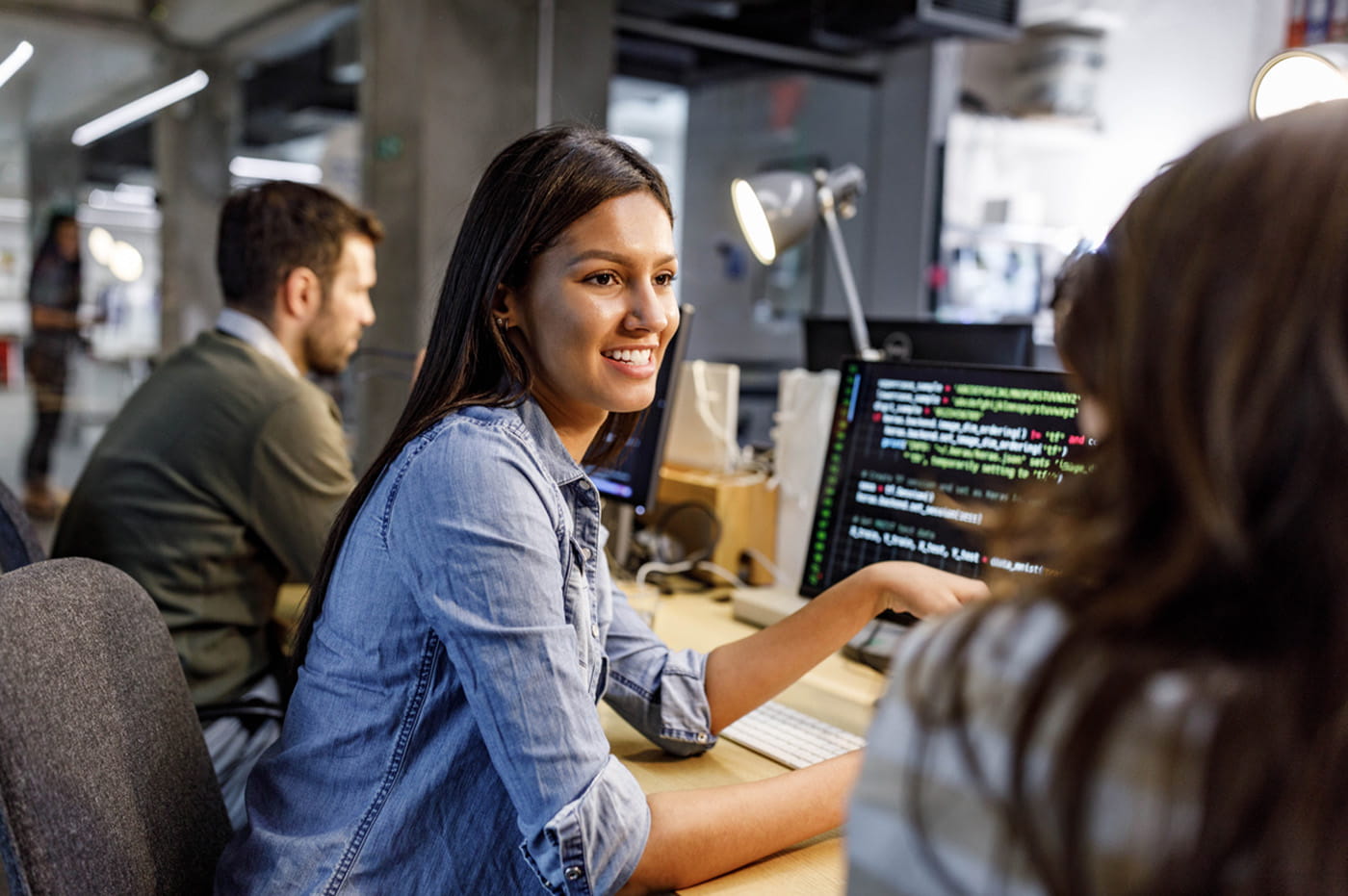 Women sitting in front of a computer with code on the screen talking to a coworker.