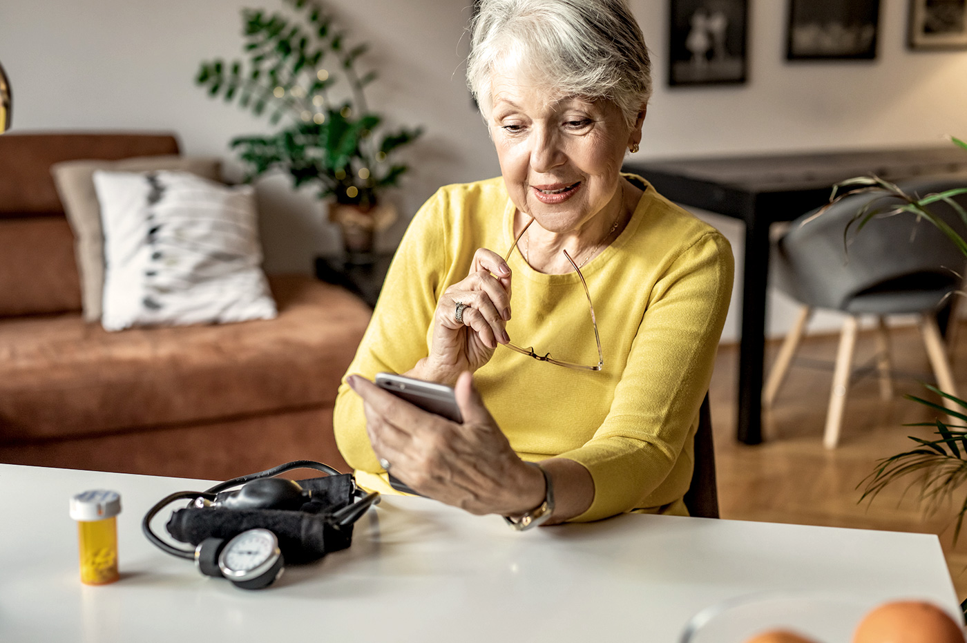 Older woman with blood pressure cuff and pill bottle on table in front of her looking at her cellphone. 