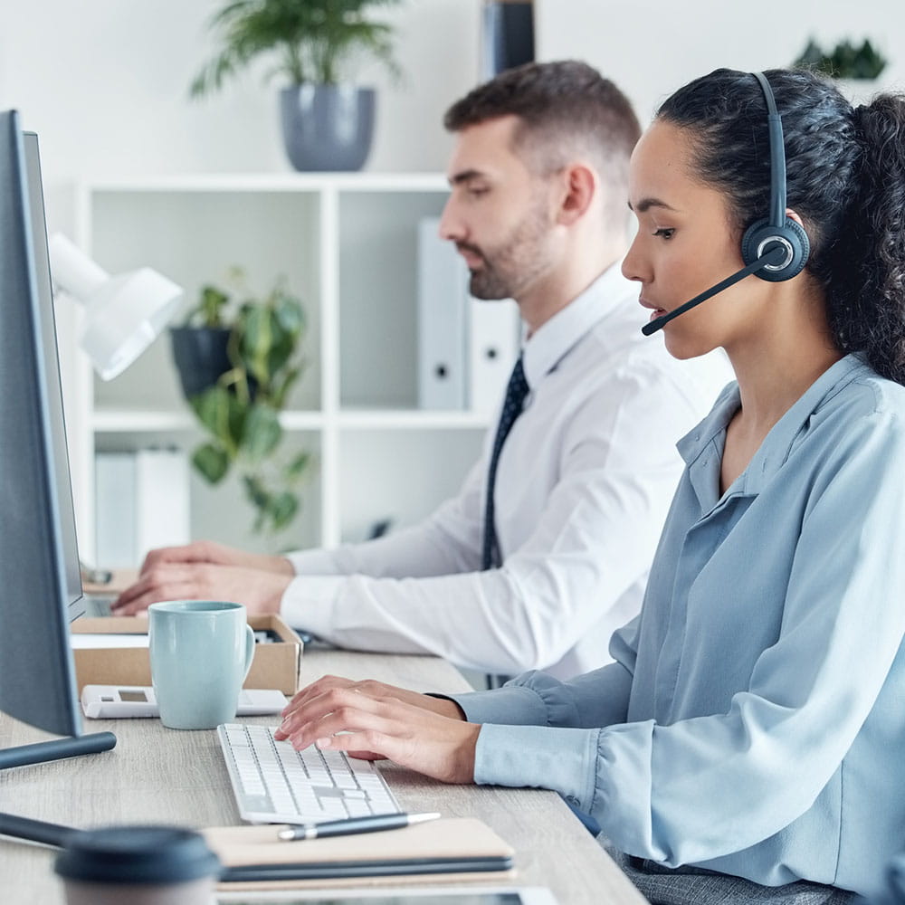 Man and Woman at work both working on computers. The Woman is wearing a headset. 