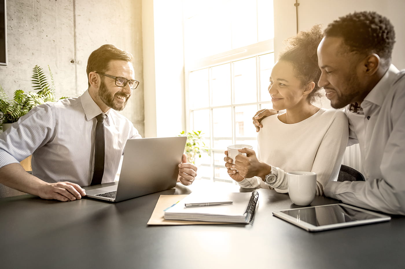 Business man talking to younger couple with laptop, iPad, and notebooks on table.