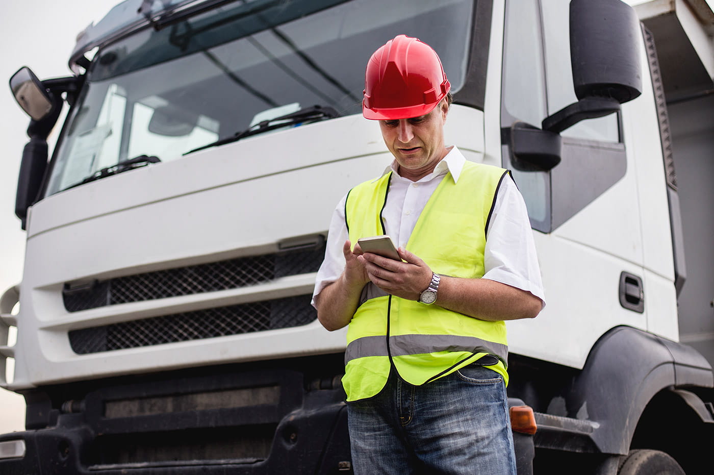 A man in a hardhat and safety vest looking at something on his cellphone.